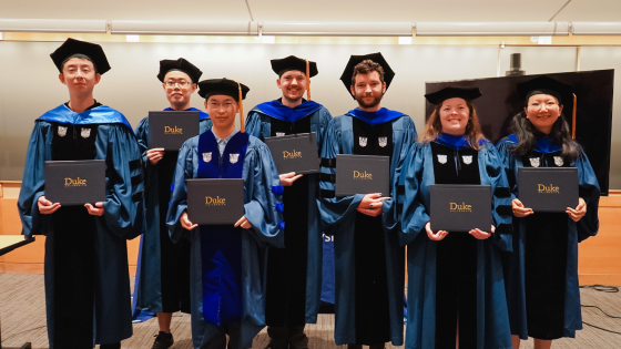 7 hooded Ph.D. graduates holding their diplomas