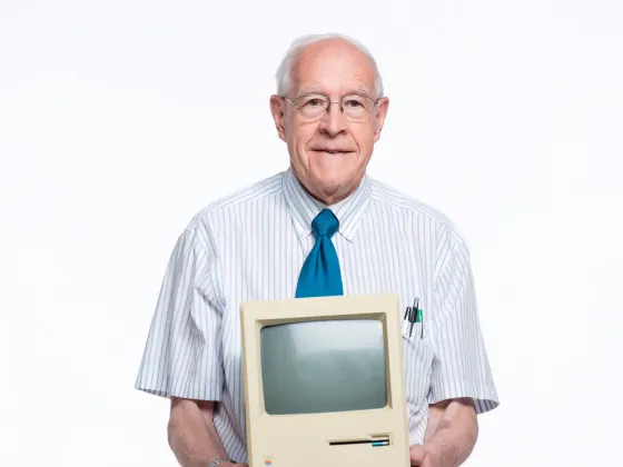Fred Brooks looks at the camera while holding an old computer with both of his hands.