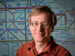 Headshot of Dr Beratan, man with glasses, wearing orange shirt, standing in front of white board